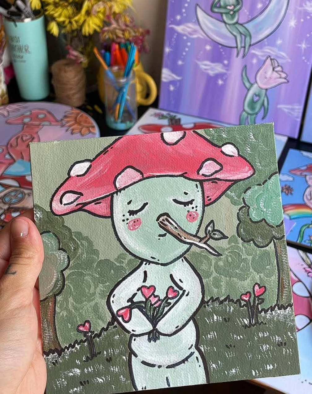 Hand holding a painting of a mushroom character carrying flowers